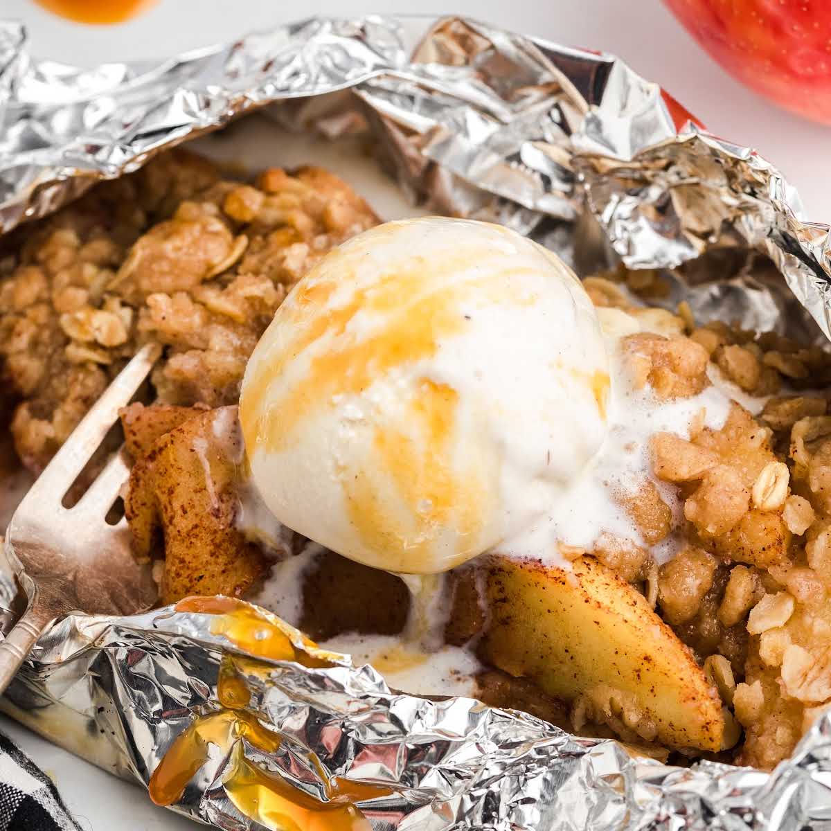Foil packet of campfire apple crisp topped with a scoop of melting vanilla ice cream and a fork beside it