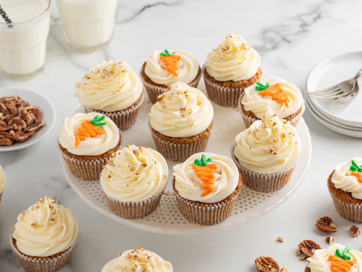 side view of carrot cake cupcakes with cream cheese frosting on a dessert stand. Some of the cupcakes are decorated with notes, some have little colorful carrots on top