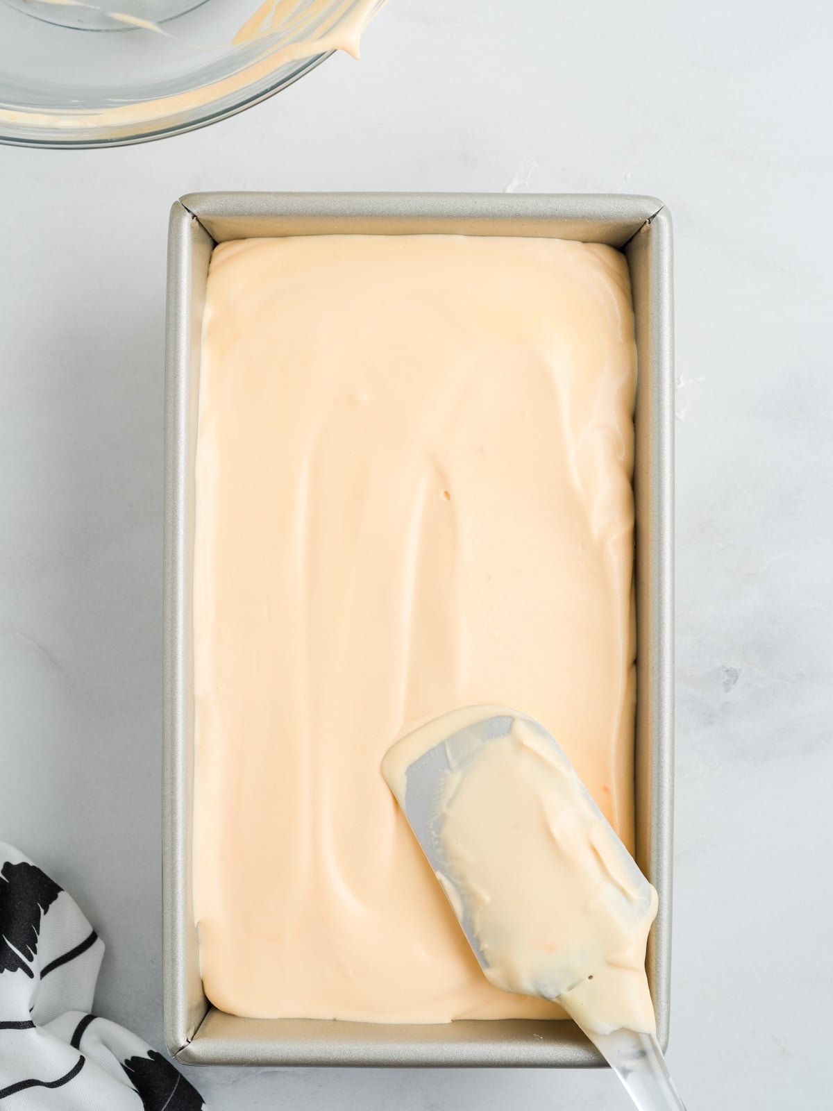 A rectangular metal loaf pan filled with smooth, creamy Cheese Ice Cream mixture being spread with a spatula, next to a black and white cloth on a light surface.