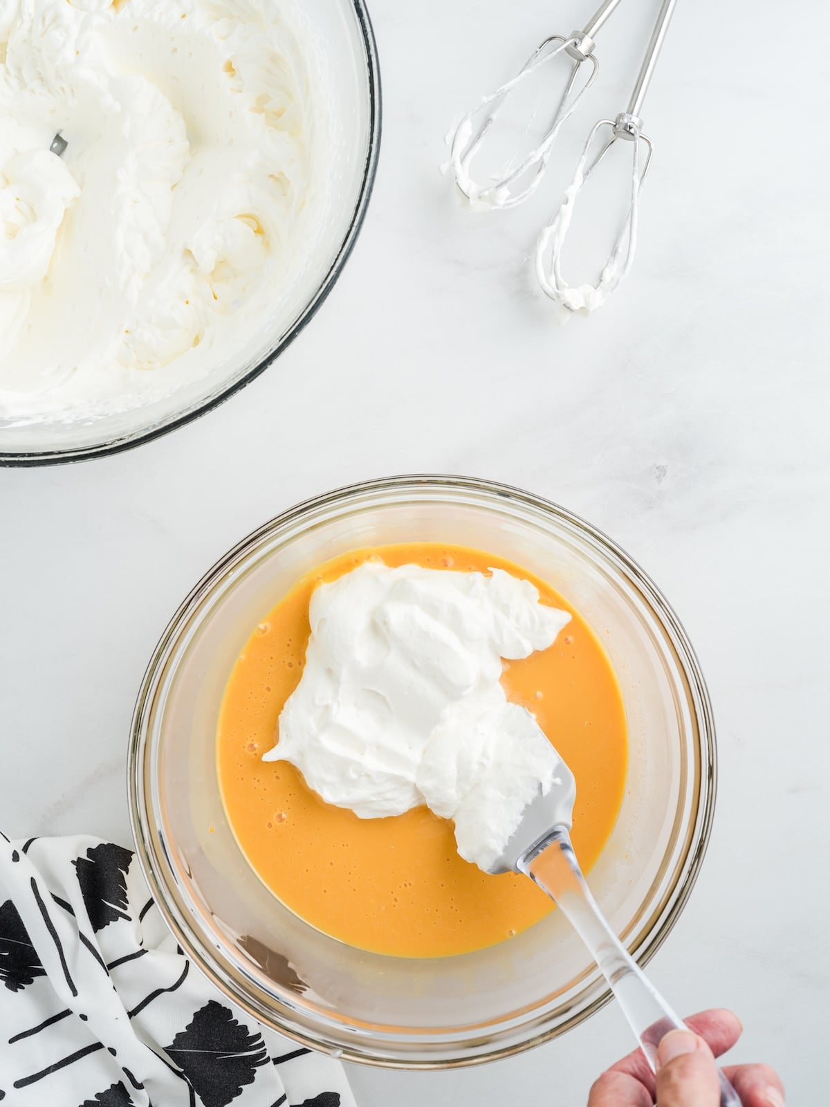A hand uses a spatula to fold whipped cream into a bowl of yellow mixture for a Cheese Ice Cream Recipe. Nearby are a bowl of whipped cream, electric beaters with some cream on them, and a white cloth with black leaf patterns.