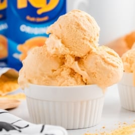 A white ramekin filled with three scoops of creamy orange-colored cheese ice cream, slightly melting. There are orange powder and another ramekin in the background, along with a blue powdered drink mix container. Perfect for a unique Cheese Ice Cream Recipe.
