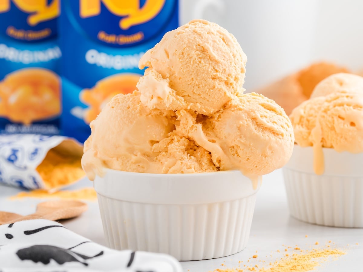 A white ramekin filled with three scoops of creamy orange-colored cheese ice cream, slightly melting. There are orange powder and another ramekin in the background, along with a blue powdered drink mix container. Perfect for a unique Cheese Ice Cream Recipe.