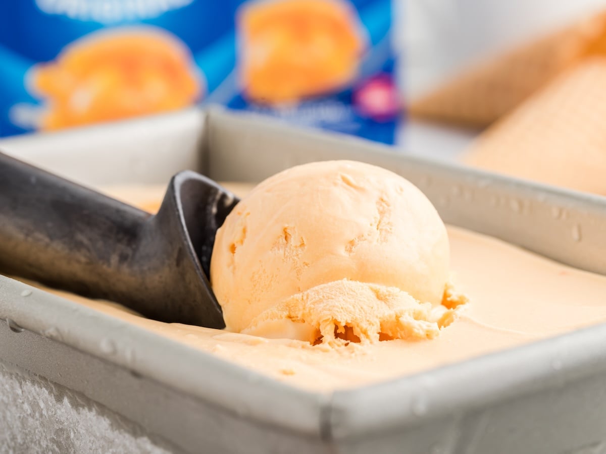 A close-up of an ice cream scoop holding a round scoop of orange sherbet from a rectangular metal container, with more sherbet and an ice cream cone blurred in the background—perfect inspiration for your next Cheese Ice Cream Recipe.