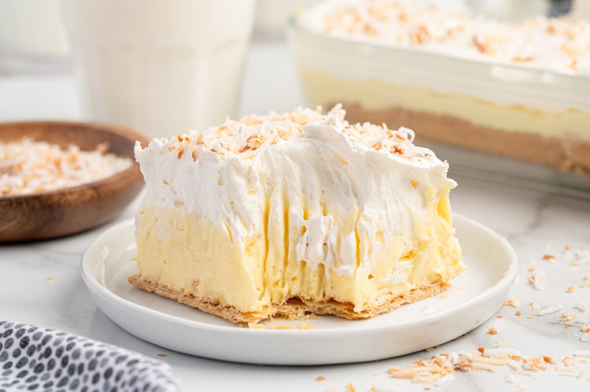 A slice of creamy coconut pie, inspired by our Coconut Cream Bars Recipe, is topped with whipped cream and toasted coconut flakes on a white plate, with more pie and toasted coconut in the background.