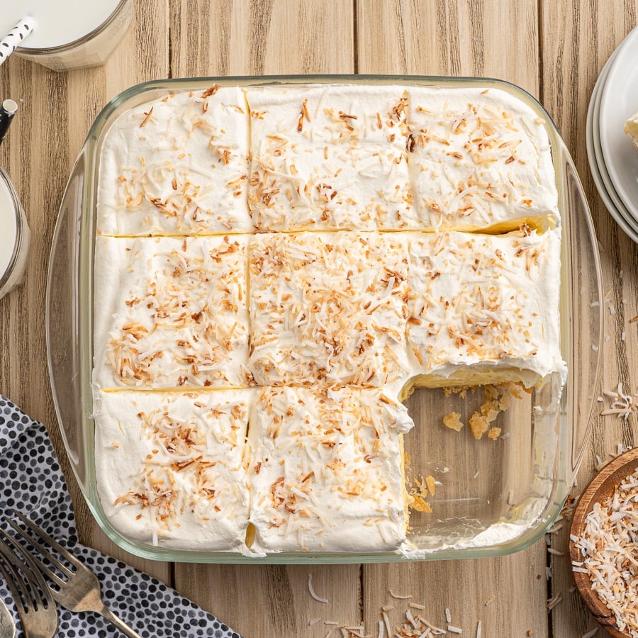 A glass baking dish filled with Coconut Cream Bars topped with whipped cream and toasted coconut flakes, cut into nine squares with one piece missing. The dish sits on a wooden surface next to plates, forks, and a bowl of coconut.