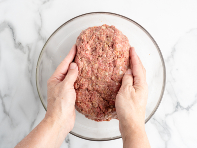 Two hands shape a large mound of raw ground meat mixed with seasonings and diced onions in a clear glass bowl, set on a white marble surface—perfect for starting your favorite meatloaf recipe.