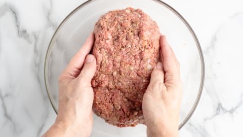Two hands shape a large mound of raw ground meat mixed with seasonings and diced onions in a clear glass bowl, set on a white marble surface&mdash;perfect for starting your favorite meatloaf recipe.