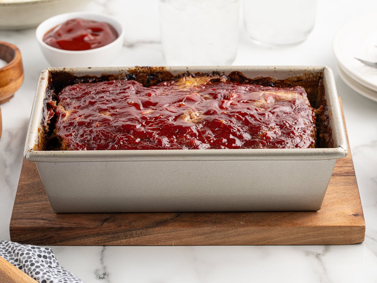 A rectangular meatloaf, made from a classic meatloaf recipe, sits in a loaf pan on a wooden board, topped with a shiny ketchup glaze. In the background, there&rsquo;s a bowl of ketchup and plates ready for serving.