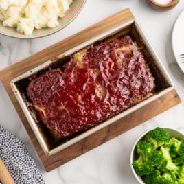 A glazed meatloaf in a loaf pan sits on a wooden board, surrounded by bowls of mashed potatoes and broccoli. A wooden spoon, a plate with forks, and a small bowl of sauce complete this classic Meatloaf Recipe scene on a marble surface.