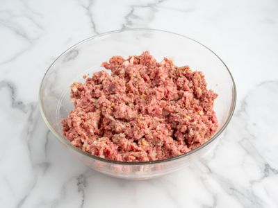 A glass bowl filled with a mixture of raw ground meat, chopped onions, and seasonings sits on a white marble surface—perfect for starting your favorite meatloaf recipe.