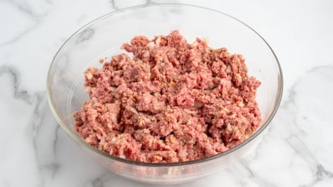A glass bowl filled with a mixture of raw ground meat, chopped onions, and seasonings sits on a white marble surface&mdash;perfect for starting your favorite meatloaf recipe.