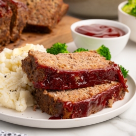 Two slices of glazed meatloaf, made from a classic meatloaf recipe, are served with mashed potatoes, broccoli florets, and a side of ketchup on a white plate. A loaf of meatloaf and a bowl of broccoli appear in the background.