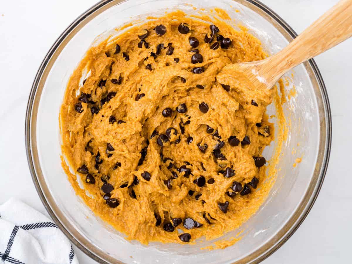 A glass bowl filled with pumpkin cookie dough mixed with chocolate chips, ready for baking delicious Pumpkin Chocolate Chip Cookies. A wooden spoon is partially submerged in the dough, and a white towel with black stripes is nearby, perfectly setting the scene to follow your favorite Pumpkin Chocolate Chip Cookies Recipe.