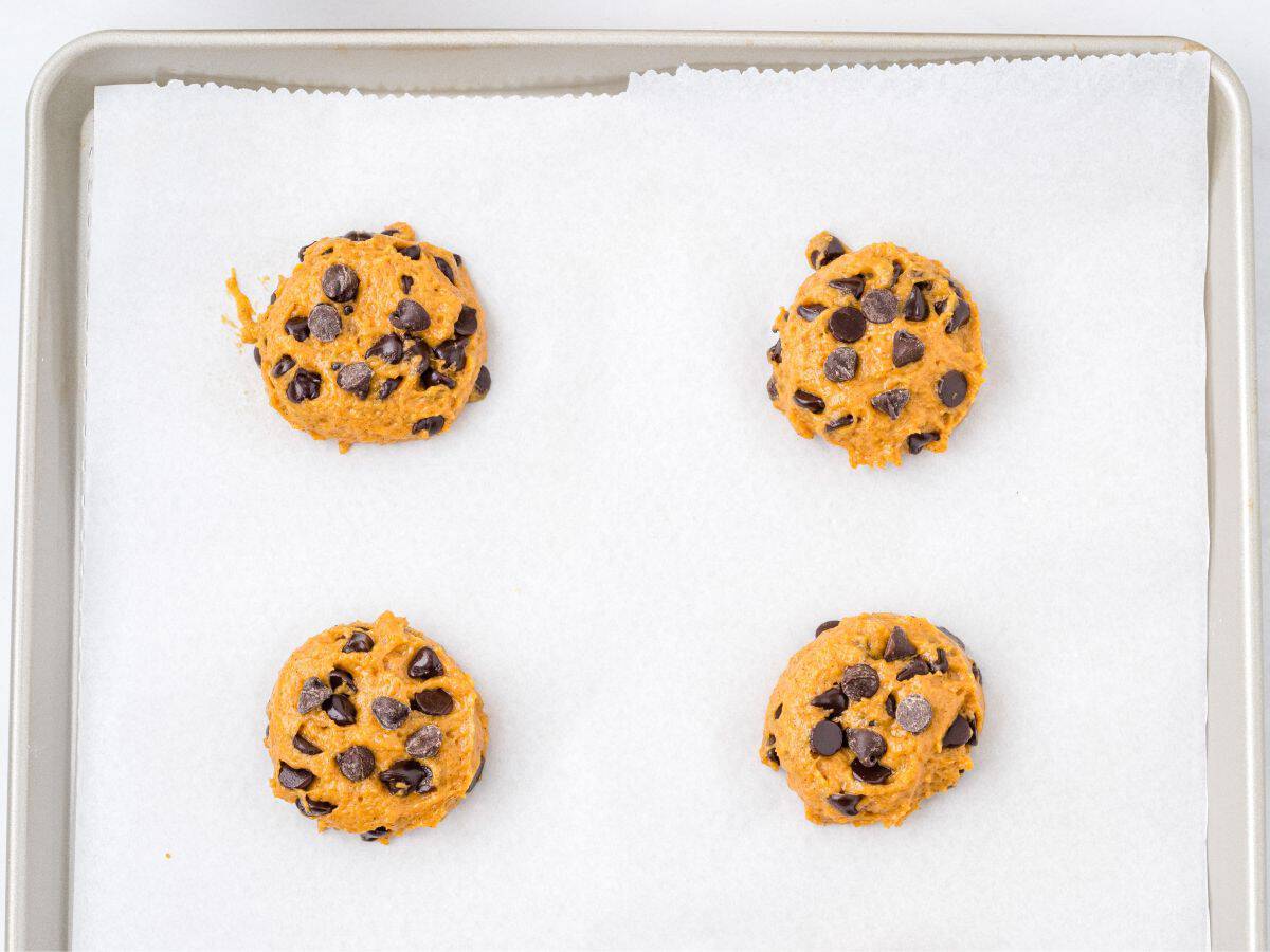 Four scoops of chocolate chip cookie dough sit spaced apart on a baking sheet lined with white parchment paper, just like in our favorite Pumpkin Chocolate Chip Cookies Recipe, ready to be baked.
