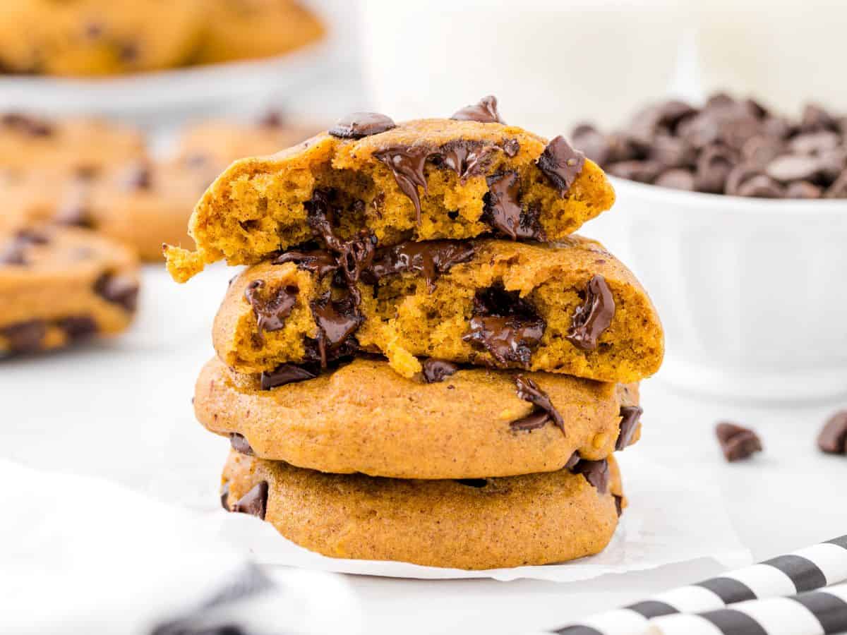 Three chocolate chip cookies are stacked, with the top cookie split in half, revealing melted chocolate inside. Reminiscent of a classic Pumpkin Chocolate Chip Cookies recipe, a bowl of chocolate chips and more cookies are blurred in the background.