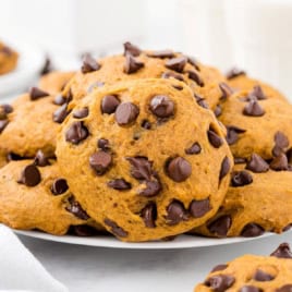 A plate piled with Pumpkin Chocolate Chip Cookies, featuring visible chocolate chips, sits on a white surface with a glass of milk in the background—an irresistible display inspired by our favorite Pumpkin Chocolate Chip Cookies Recipe.