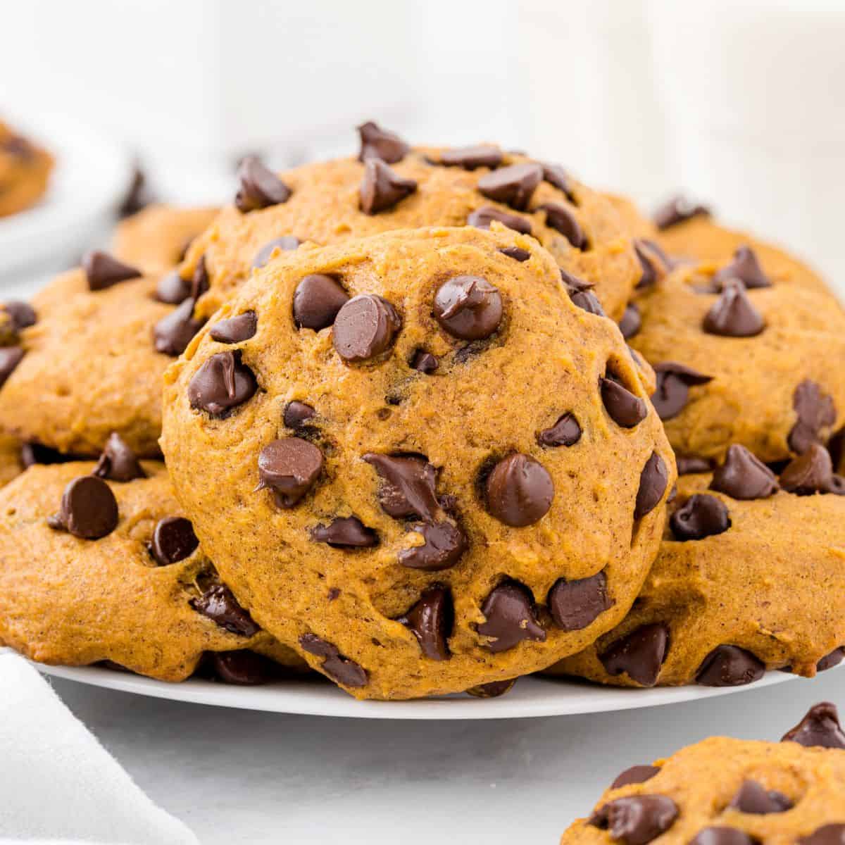 A plate piled with Pumpkin Chocolate Chip Cookies, featuring visible chocolate chips, sits on a white surface with a glass of milk in the background—an irresistible display inspired by our favorite Pumpkin Chocolate Chip Cookies Recipe.