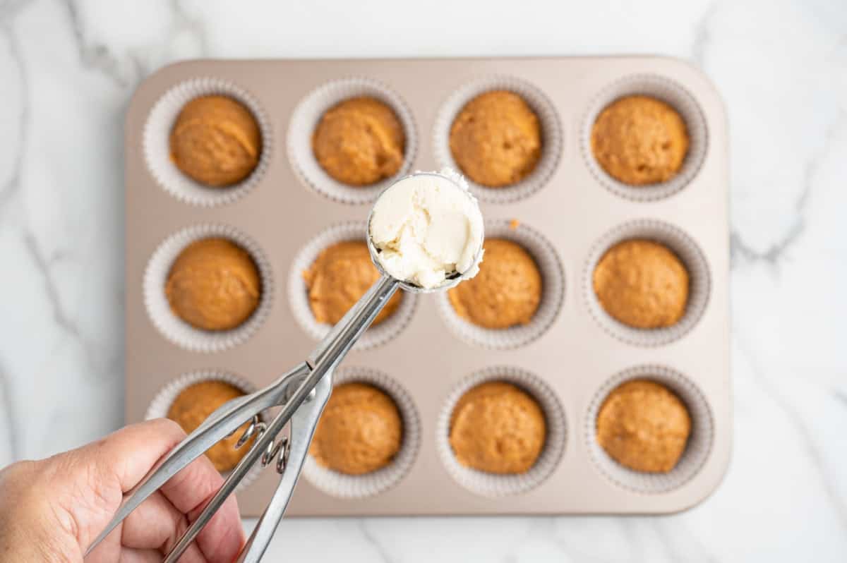 A hand holds an ice cream scoop with cream cheese filling above a muffin tin lined with paper cups, some filled with batter, on a marble surface—perfectly capturing the process of making Pumpkin Cream Cheese Muffins or following your favorite Pumpkin Cream Cheese Muffins Recipe.