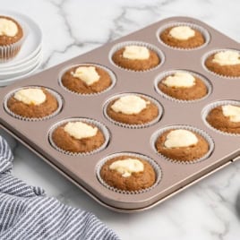 A muffin tin with twelve Pumpkin Cream Cheese Muffins, each with a luscious cream cheese center, sits on a marble counter next to a striped cloth and stacked white plates.