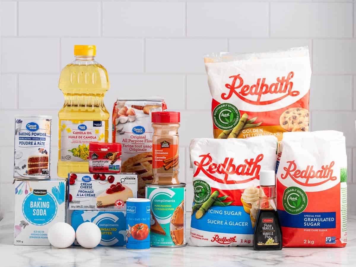 A variety of baking ingredients, including flour, sugars, oil, eggs, baking soda, baking powder, pumpkin puree, vanilla, cinnamon, salt, and other essentials needed for a delicious Pumpkin Cream Cheese Muffins Recipe are displayed on a kitchen counter.