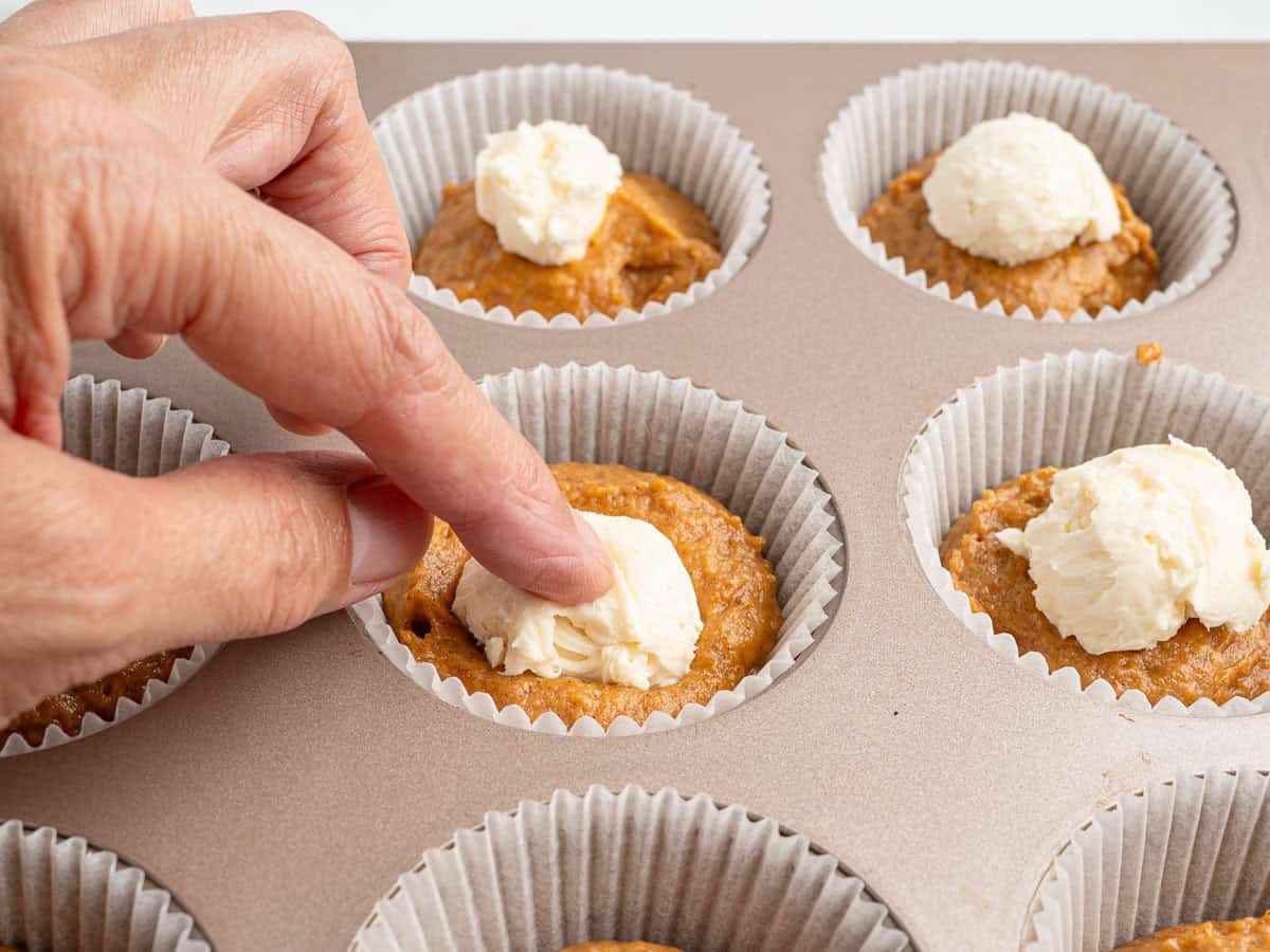 A hand presses a finger into pumpkin muffin batter in a muffin tin, creating space for a dollop of cream cheese filling in each liner—a simple step that brings these Pumpkin Cream Cheese Muffins to life.