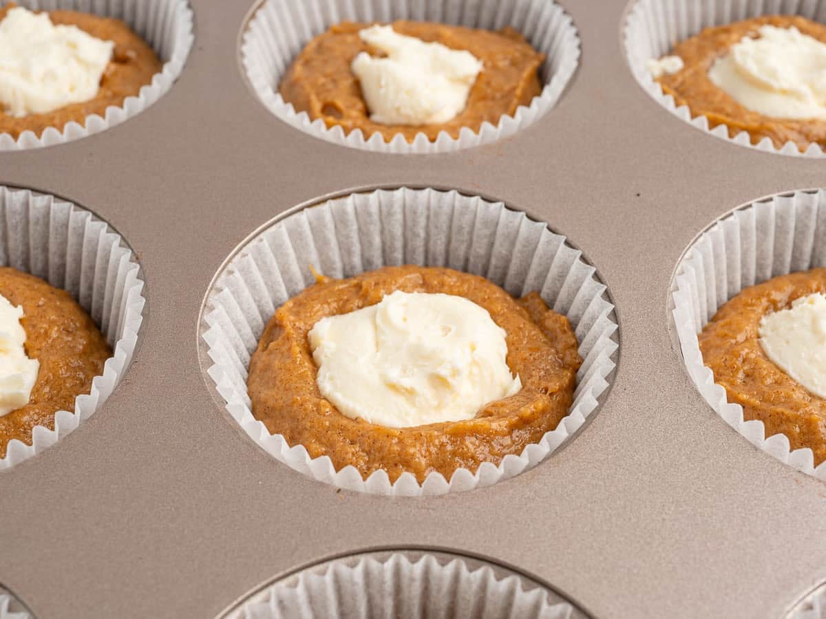 A muffin tin filled with paper liners, each holding pumpkin muffin batter topped with a dollop of cream cheese before baking—just what you'd expect when following a classic Pumpkin Cream Cheese Muffins Recipe.