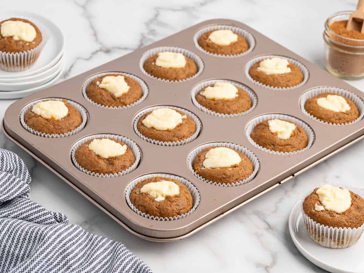 A muffin tray holds 12 freshly baked Pumpkin Cream Cheese Muffins, each with a dollop of cream cheese in the center. A single muffin sits on a plate nearby, next to a jar of cinnamon sugar and a blue-striped towel on a marble countertop.