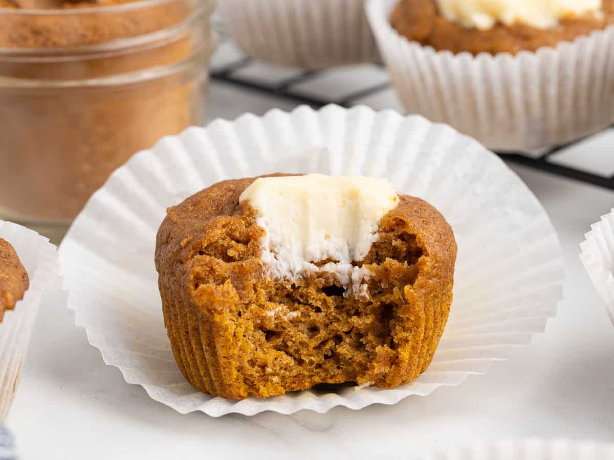 A pumpkin muffin with cream cheese filling sits in a white paper liner. The muffin has a bite taken out, revealing its moist texture and creamy center—the hallmark of an irresistible Pumpkin Cream Cheese Muffin. Other muffins and a glass jar are blurred in the background, capturing the cozy essence of this classic Pumpkin Cream Cheese Muffins recipe.