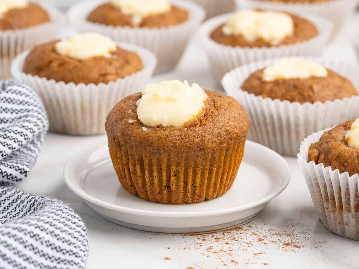 A pumpkin muffin with cream cheese filling sits on a white plate, surrounded by more Pumpkin Cream Cheese Muffins in paper liners. A striped towel is visible on the side, inviting you to try this irresistible Pumpkin Cream Cheese Muffins Recipe.