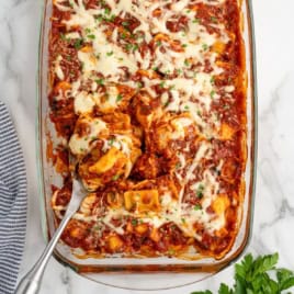A glass baking dish filled with cheesy baked tortellini in tomato sauce, topped with melted cheese and parsley, offers a cozy twist reminiscent of a Ravioli Lasagna Recipe. A serving spoon is lifting a portion from the dish, revealing layers of gooey goodness. A striped towel and fresh parsley are nearby.