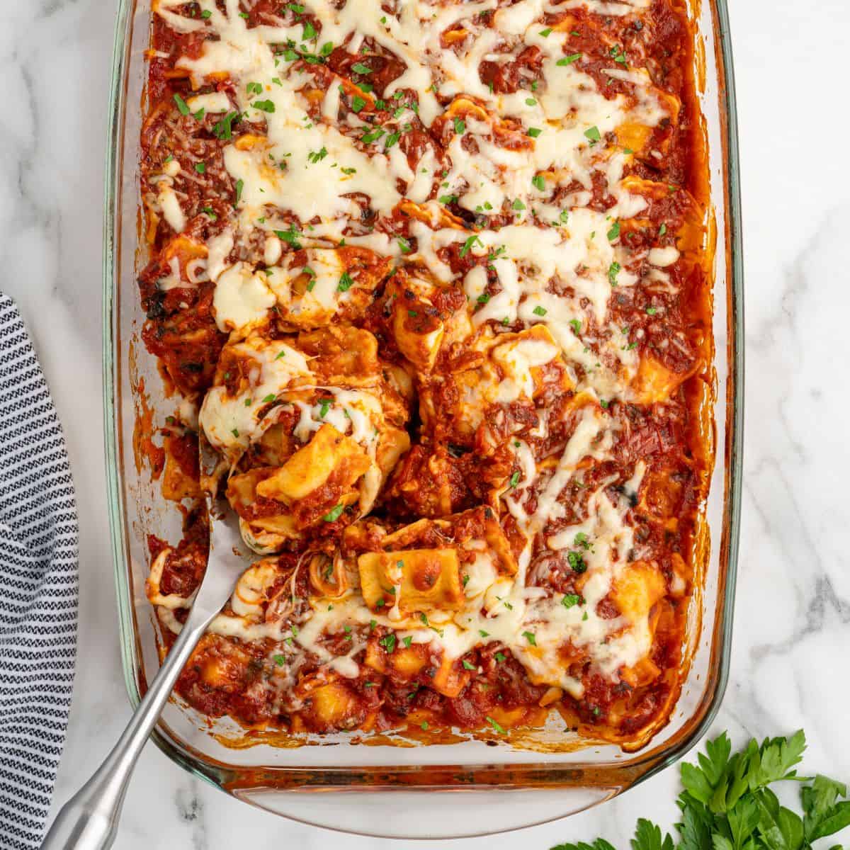 A glass baking dish filled with cheesy baked tortellini in tomato sauce, topped with melted cheese and parsley, offers a cozy twist reminiscent of a Ravioli Lasagna Recipe. A serving spoon is lifting a portion from the dish, revealing layers of gooey goodness. A striped towel and fresh parsley are nearby.