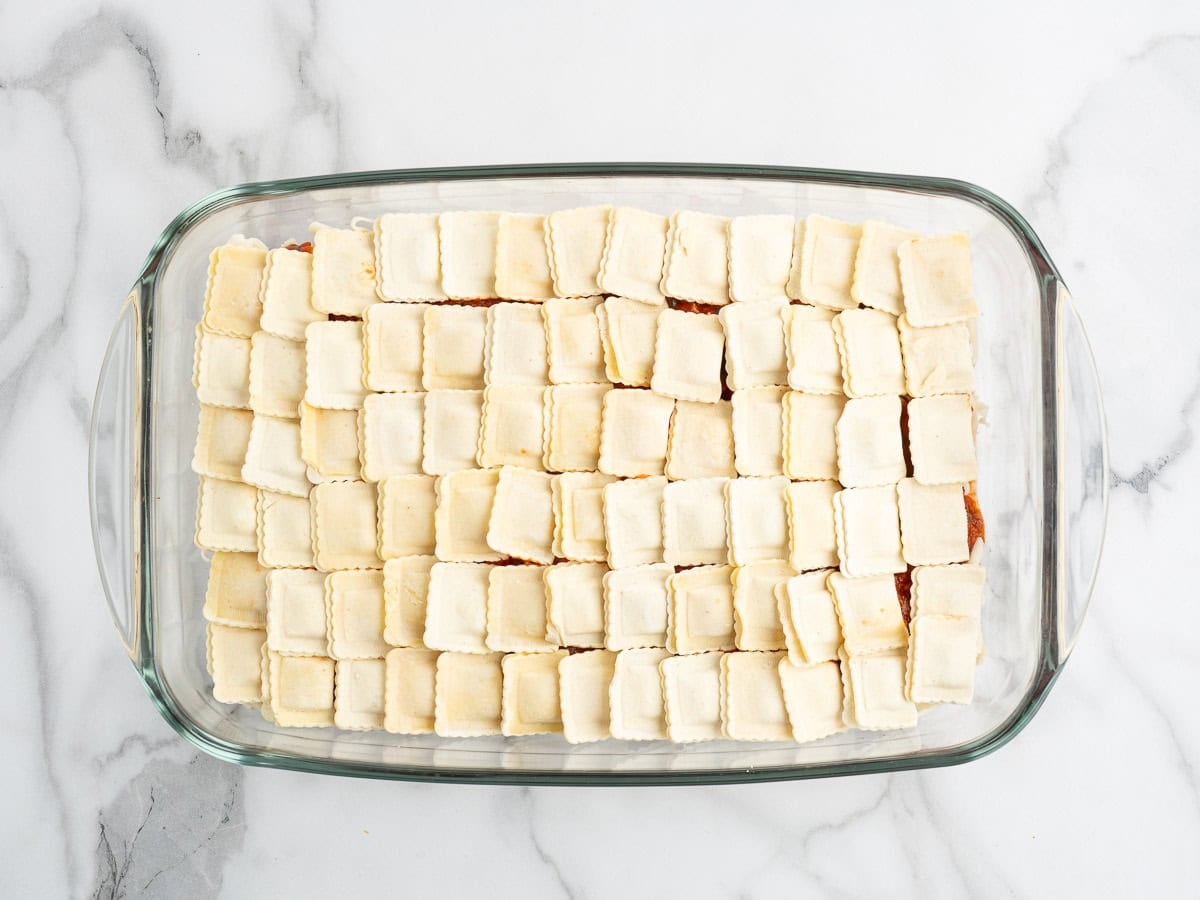 A glass baking dish filled with neatly arranged rows of uncooked square ravioli on a white marble surface, ready to be transformed into a delicious Ravioli Lasagna Recipe.