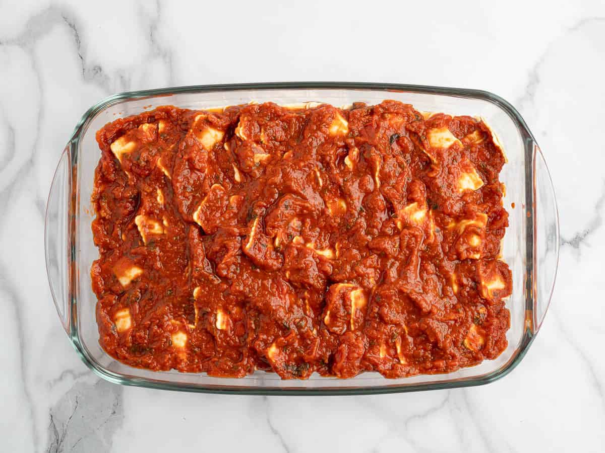 A glass baking dish filled with Ravioli Lasagna, topped with a thick layer of tomato sauce and visible chunks of cheese, sits on a white marble surface—an inviting take on the classic Ravioli Lasagna Recipe.