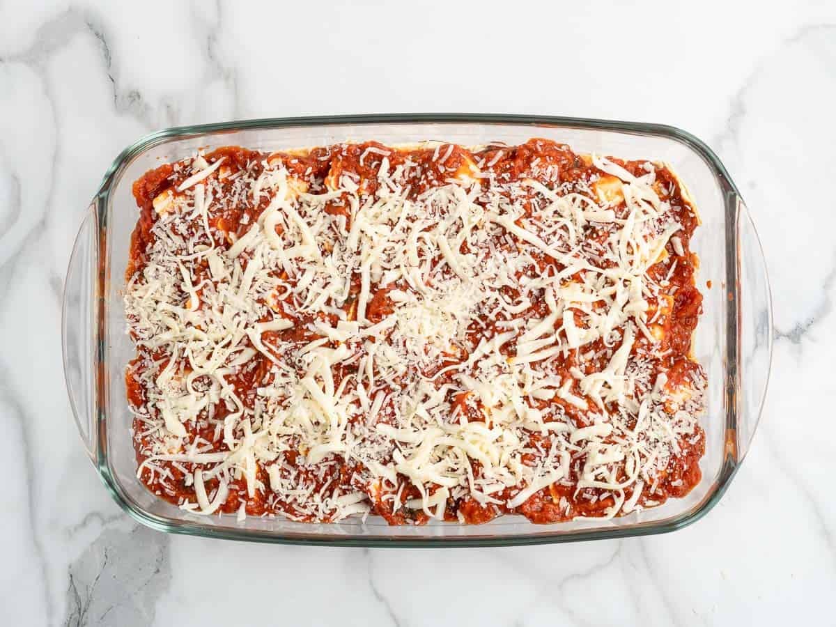 A glass baking dish filled with Ravioli Lasagna, topped with tomato sauce and shredded cheese, sits on a marble countertop, ready to be baked—a delicious twist inspired by the classic Ravioli Lasagna Recipe.
