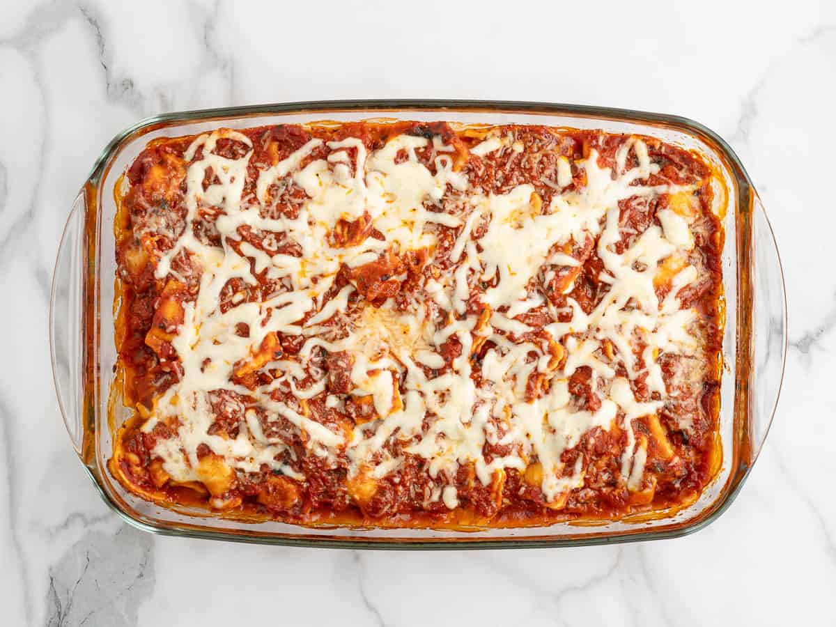A rectangular glass dish filled with baked Ravioli Lasagna, topped with melted cheese and tomato sauce, sits on a white marble surface—an enticing take on the classic Ravioli Lasagna Recipe.