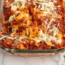 A glass baking dish filled with a delicious Ravioli Lasagna bake in tomato sauce, topped with melted cheese and chopped herbs. A serving spoon is lifting a portion out, showcasing this easy Ravioli Lasagna Recipe.