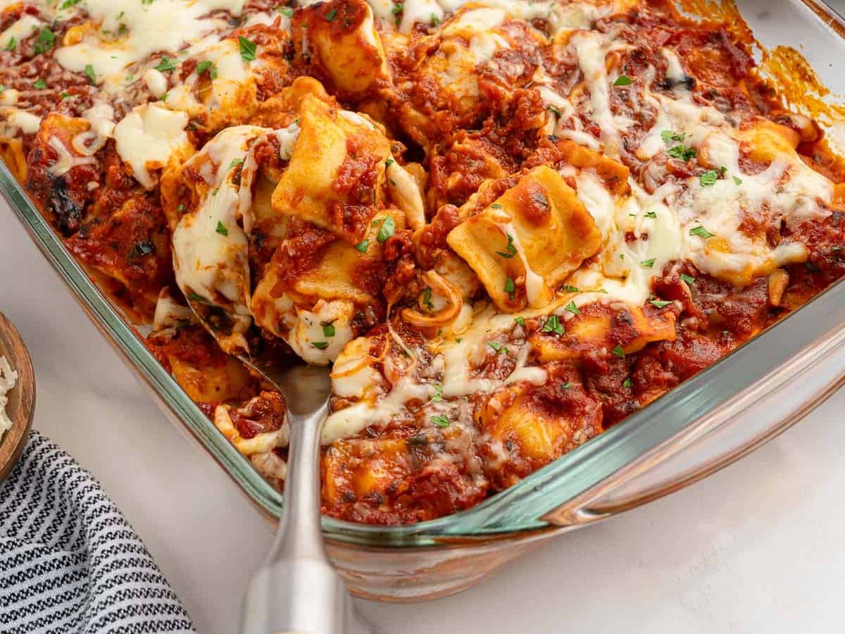 A glass baking dish filled with cheesy baked Ravioli Lasagna topped with tomato sauce and melted mozzarella, garnished with chopped parsley. A serving spoon is scooping out a portion, showcasing this delicious Ravioli Lasagna Recipe.