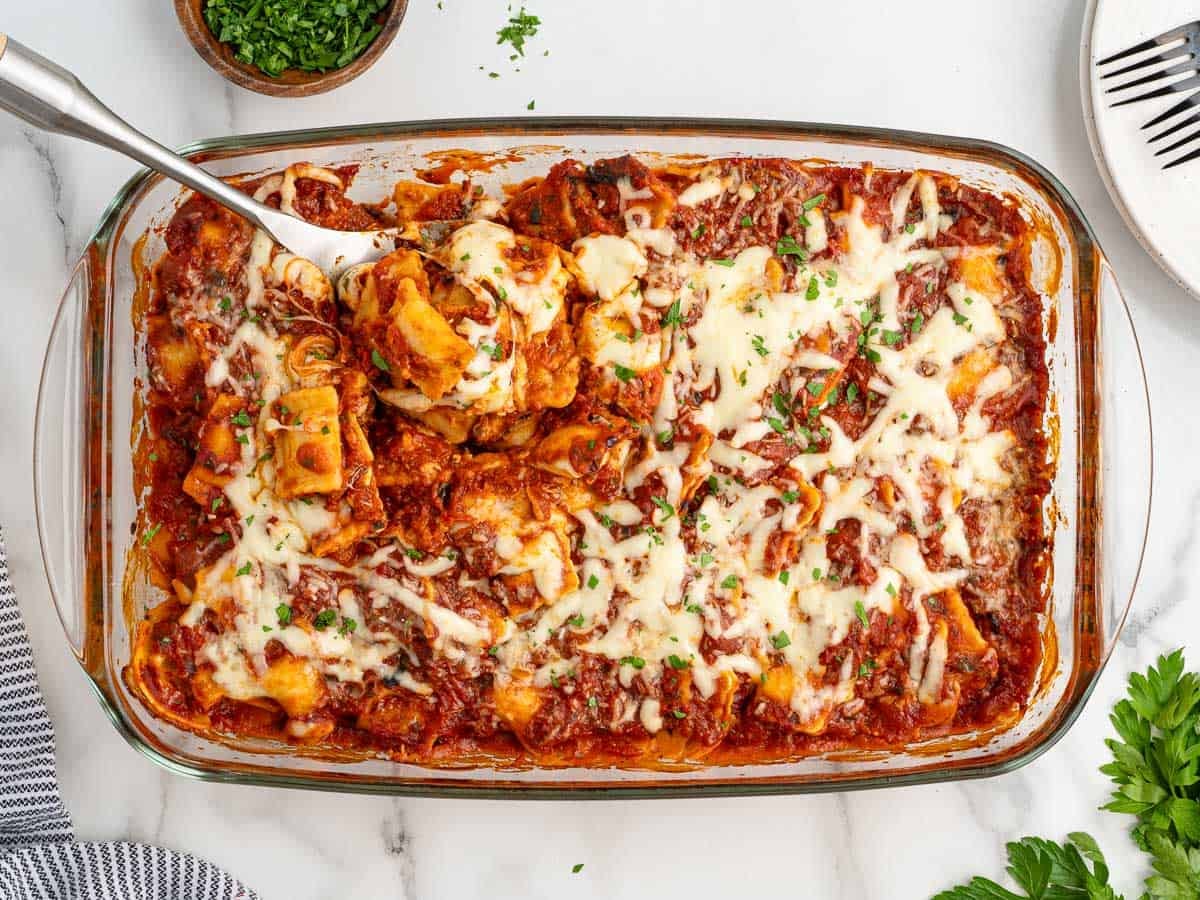 A glass baking dish filled with cheesy baked pasta in tomato sauce, this Ravioli Lasagna is topped with melted cheese and chopped herbs. A serving spoon is lifting out a portion of this irresistible Ravioli Lasagna Recipe. Plates, utensils, and herbs are nearby on a marble surface.
