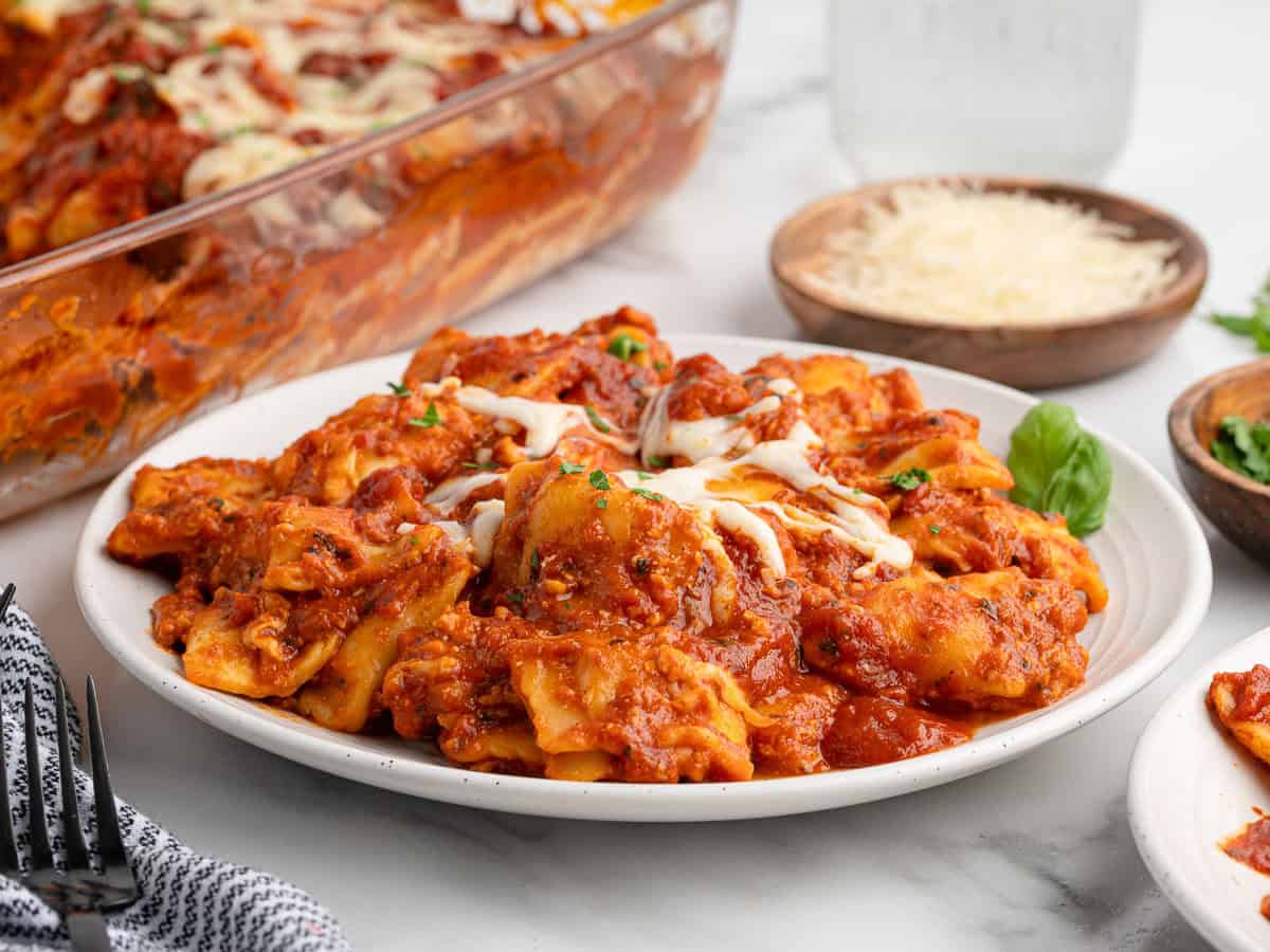 A plate of baked ravioli covered in tomato sauce and melted cheese sits on a table, alongside a baking dish filled with savory Ravioli Lasagna and bowls of shredded cheese nearby.