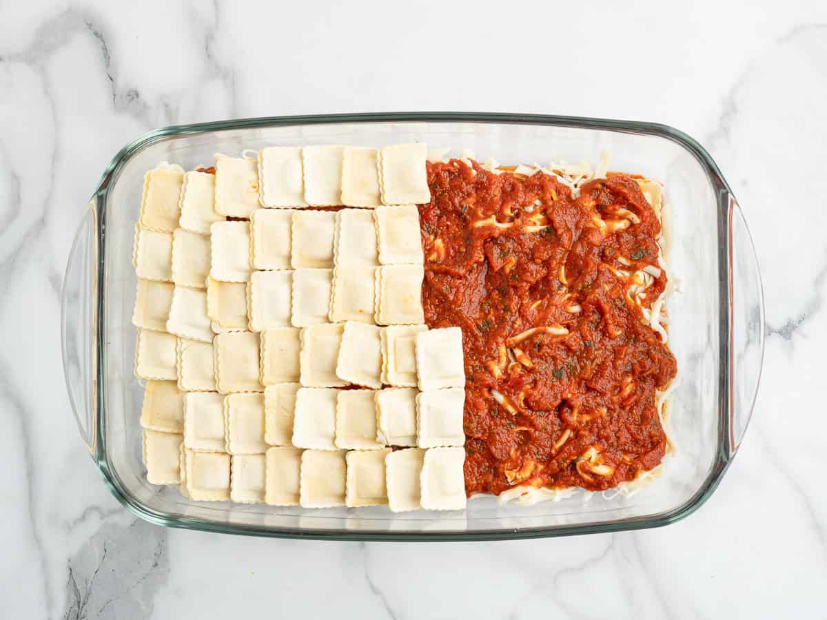 A glass baking dish with one half filled with uncooked ravioli and the other half topped with red tomato sauce and shredded cheese, on a marble surface—perfectly showcasing this easy Ravioli Lasagna Recipe.