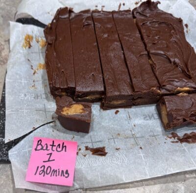 Chocolate Peanut Butter Fudge bars with gorgeous swirls of creamy peanut butter are cut on parchment paper, ready to enjoy. One delectable piece is set apart, highlighting the irresistible texture of this Chocolate Peanut Butter Fudge Recipe. A pink sticky note nearby reads Batch 1, 120 mins.