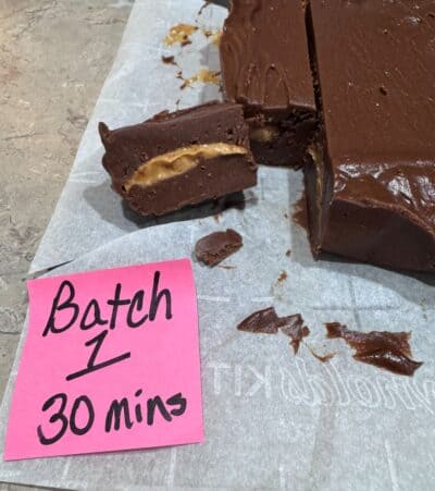 A batch of Chocolate Peanut Butter Fudge with a creamy peanut butter layer is cut into pieces on parchment paper. A pink sticky note nearby reads, Batch 1 30 mins, marking the start of this delicious Chocolate Peanut Butter Fudge Recipe.