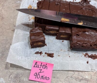 Chocolate Peanut Butter Fudge squares with a creamy peanut butter layer are partially cut on parchment paper, embodying the irresistible charm of a classic Chocolate Peanut Butter Fudge Recipe. A knife sits on top, ready for serving. A pink sticky note below reads Batch 1, 90 mins.
