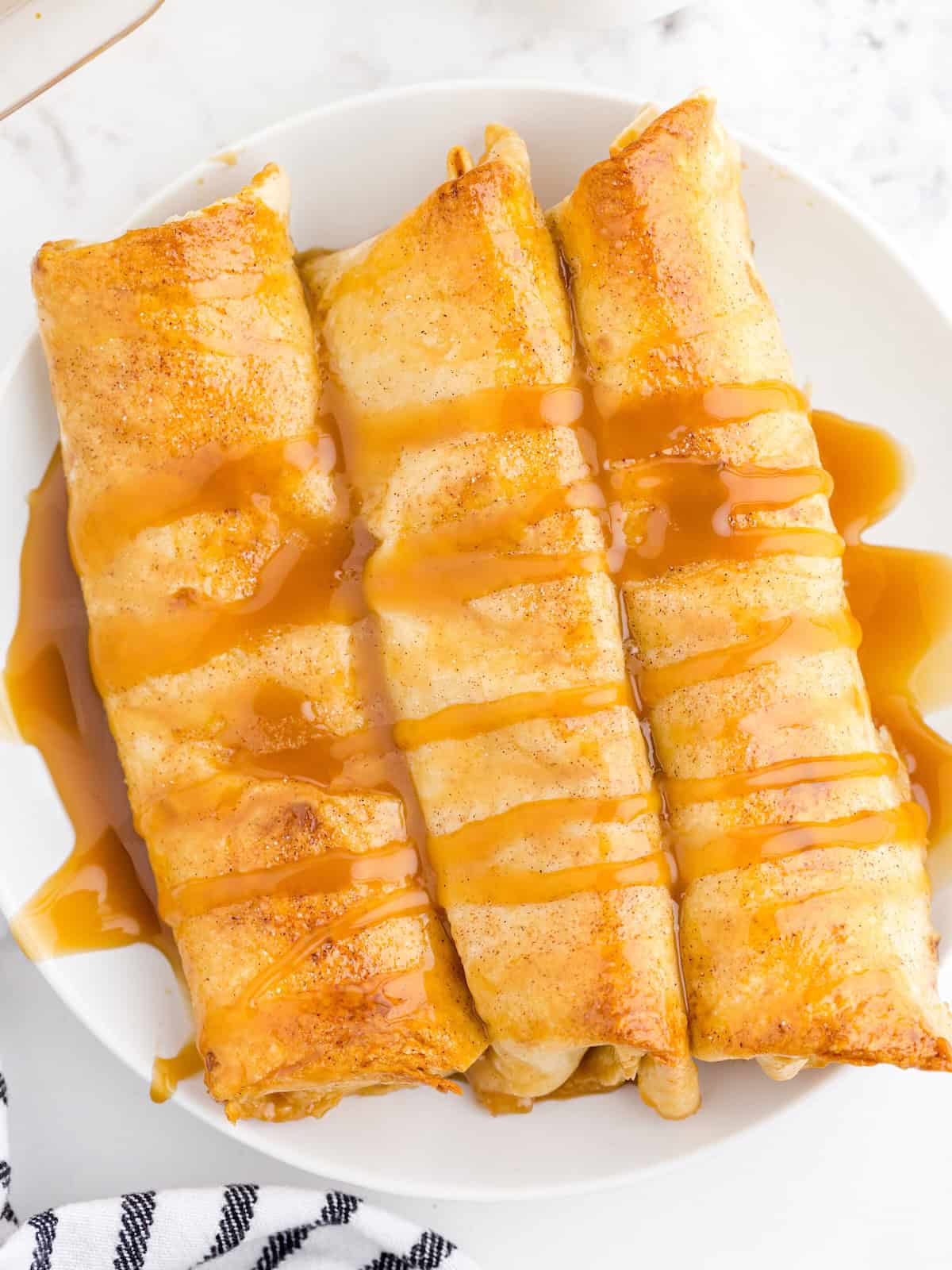 Three golden-brown baked desserts from an irresistible Apple Enchiladas Recipe are drizzled with caramel sauce and arranged side by side on a white plate, set on a light marble surface with a striped cloth partially visible at the bottom left.