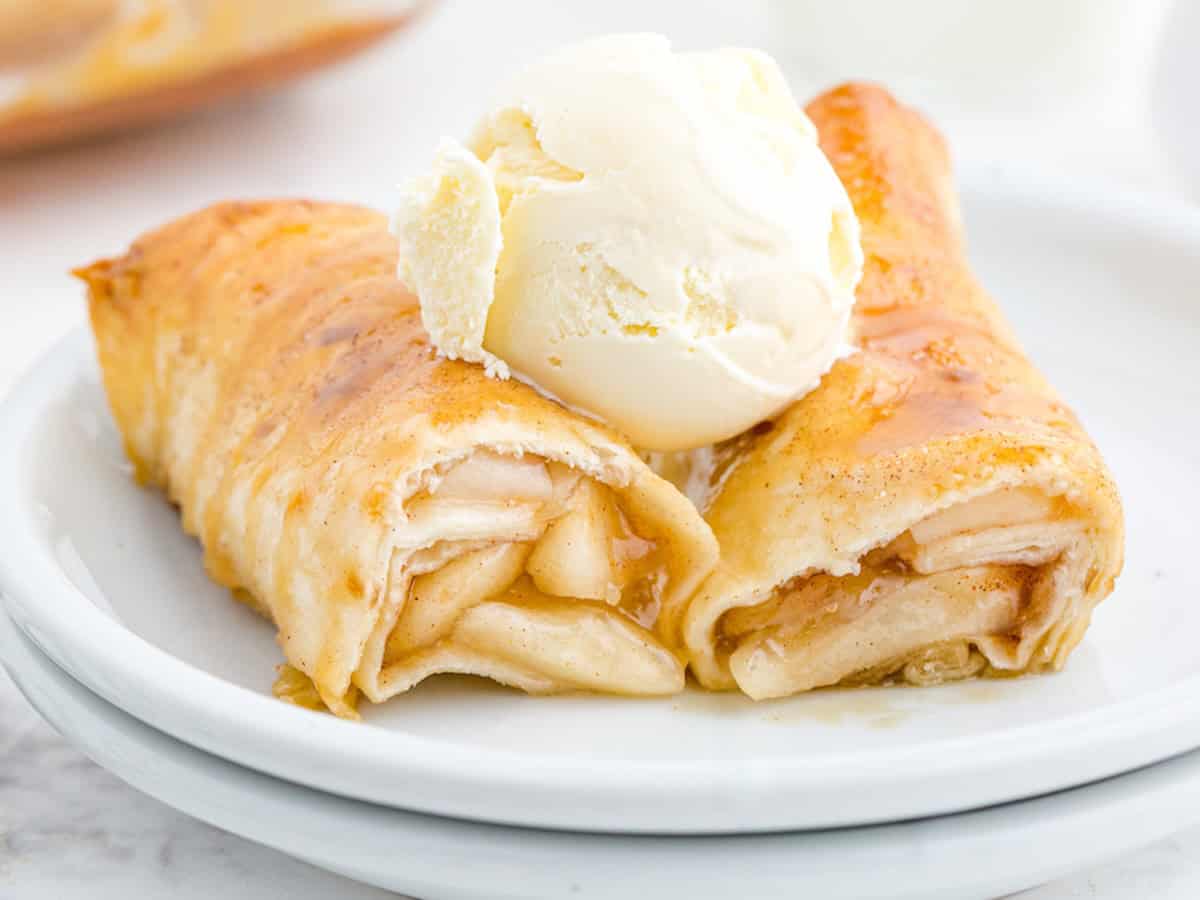 Two baked apple-filled crepes, inspired by our Apple Enchiladas Recipe, are topped with a scoop of vanilla ice cream and drizzled with syrup, served on a white plate.