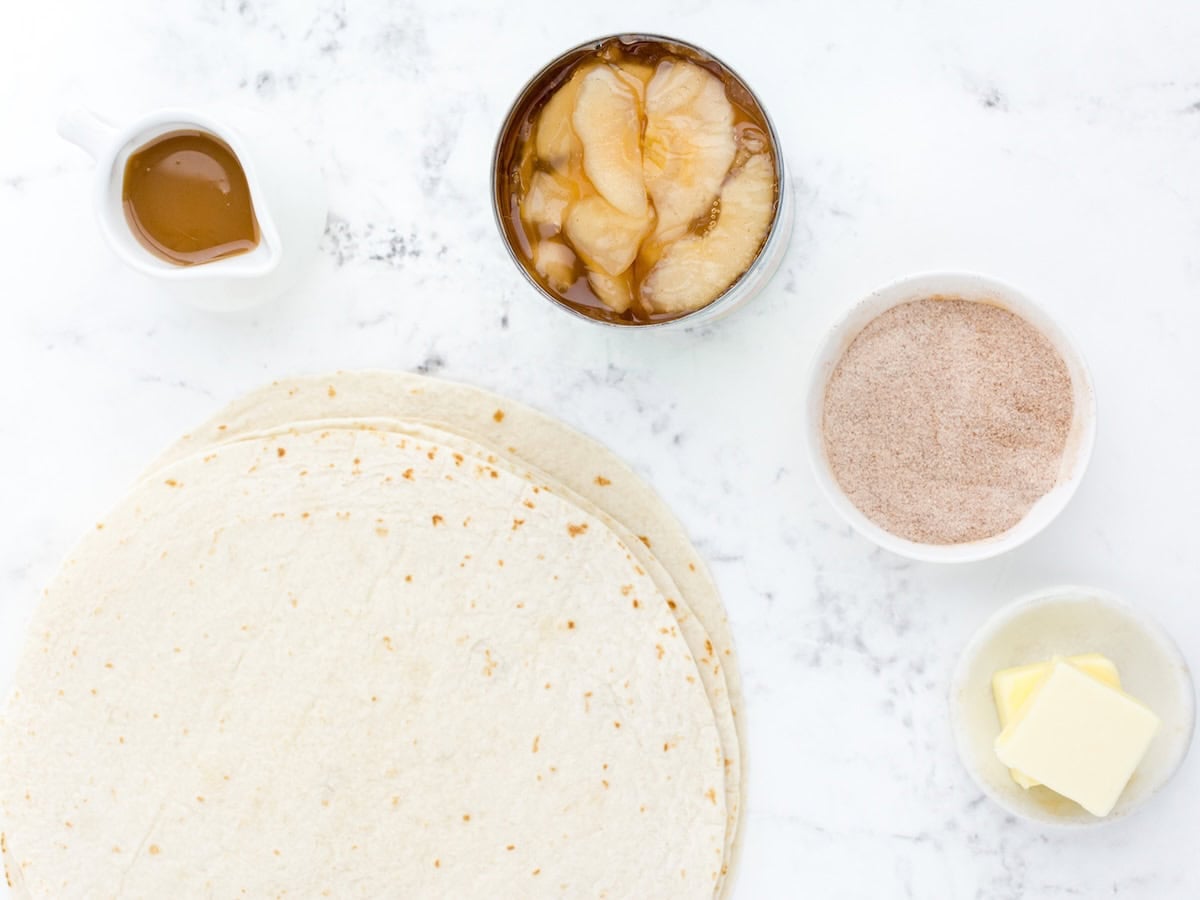 Five ingredients on a white surface set the stage for this delicious Apple Enchiladas recipe: flour tortillas, canned apple pie filling, a bowl of cinnamon sugar, a small dish of butter, and a small pitcher of caramel sauce.