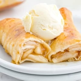 Two apple-filled crepes, reminiscent of a classic Apple Enchiladas Recipe, are topped with a scoop of vanilla ice cream and drizzled with syrup, then served elegantly on a white plate.