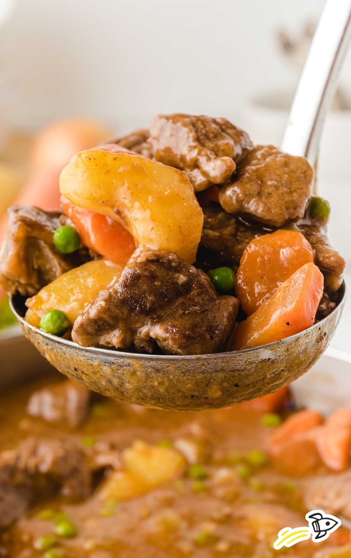 Ladle full of beef stew lifted from a pot, showing tender meat, potatoes, carrots, and peas in savory broth.