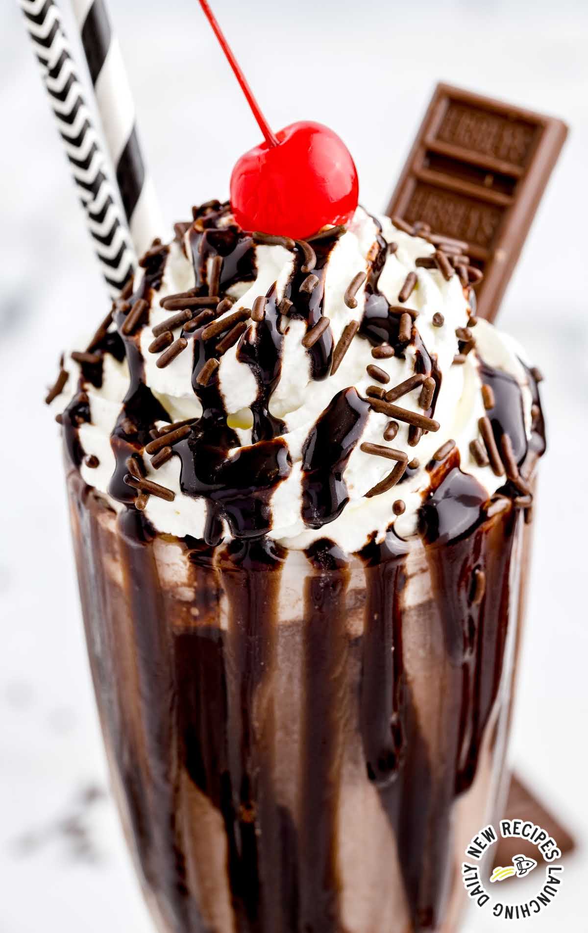 close up overhead shot of a glass of Chocolate Milkshake topped with whipped cream then garnished with Hershey bars, a cherry, chocolate sprinkles, and chocolate syrup
