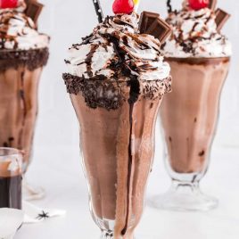 close up shot of glasses of Chocolate Milkshake topped with whipped cream then garnished with Hershey bars, a cherry, chocolate sprinkles, and chocolate syrup